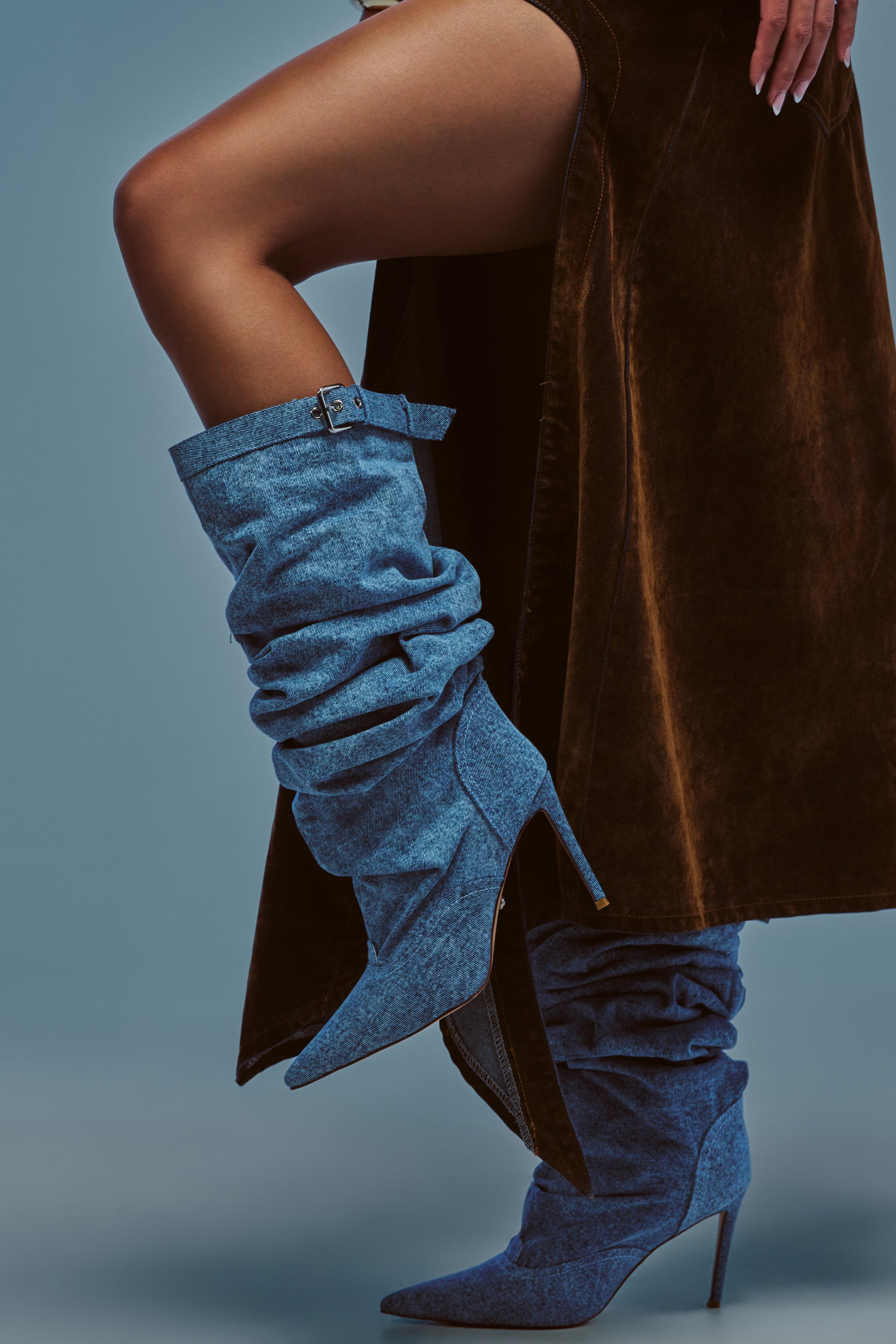 A person wearing a brown skirt and the AZALEA WANG DARIA DENIM STILETTO BOOT—with one leg raised and white-painted nails—stands against a plain blue background.