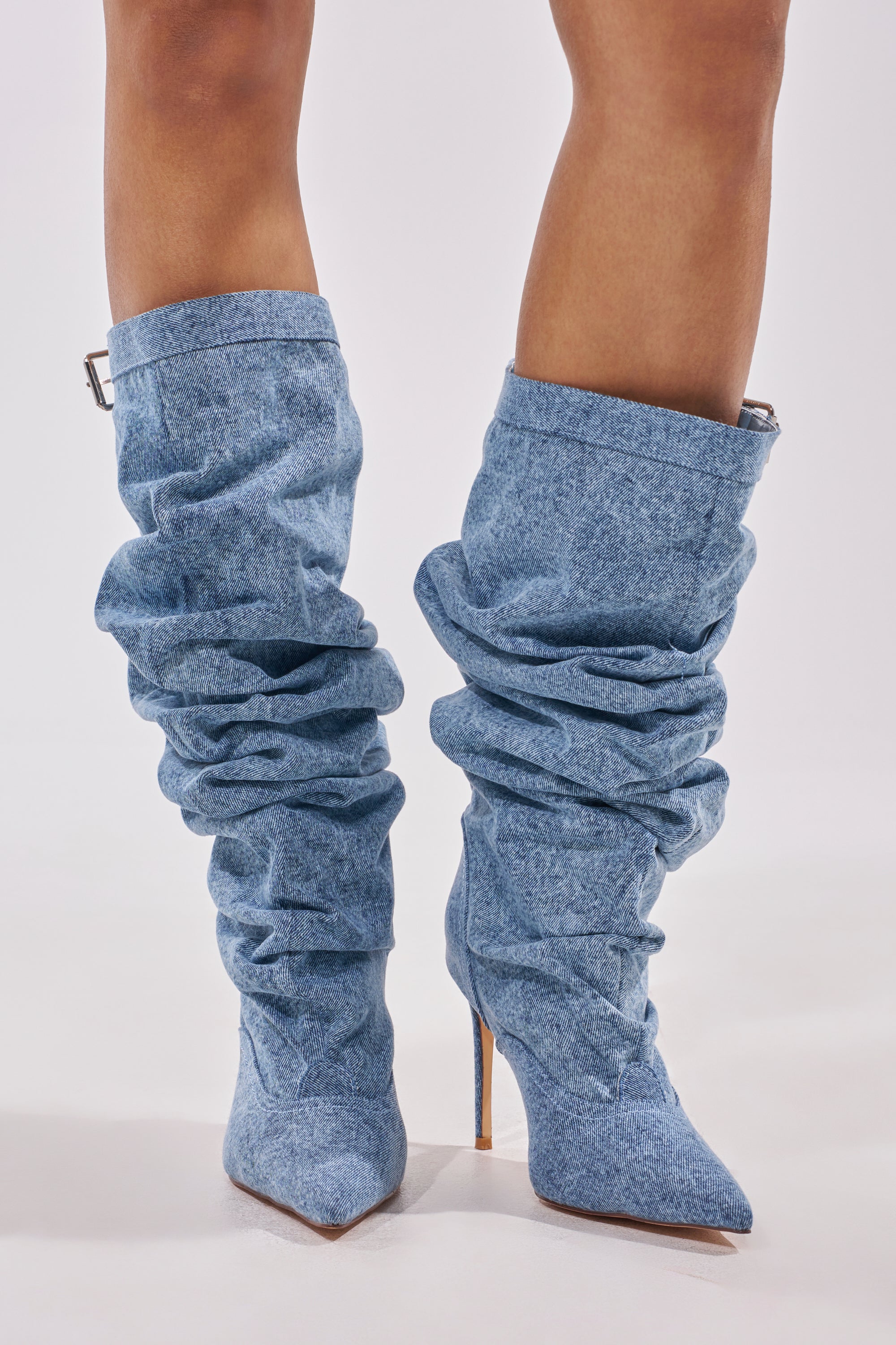 A person wears the AZALEA WANG DARIA DENIM STILETTO BOOT—light blue, slouchy, knee-high denim with pointed toes and stiletto heels—while standing against a plain white background. 1