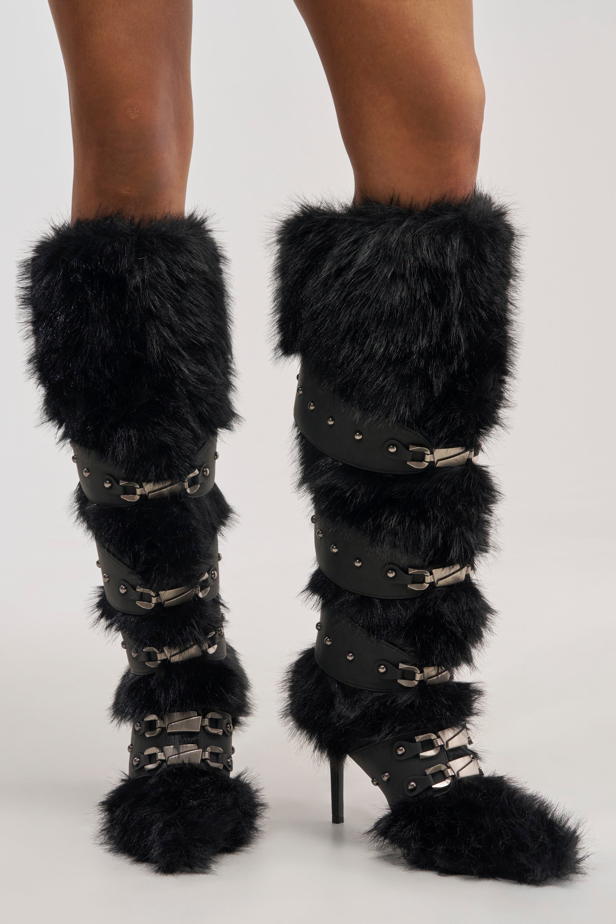 Wearing the AKIRA GOLD DARIO BLACK FUR WITH STRAPS KNEE HIGH BOOT, featuring tall black faux fur, multiple buckle straps, and studded details, this person stands against a plain background—these sexy winter boots make a bold fashion statement.
