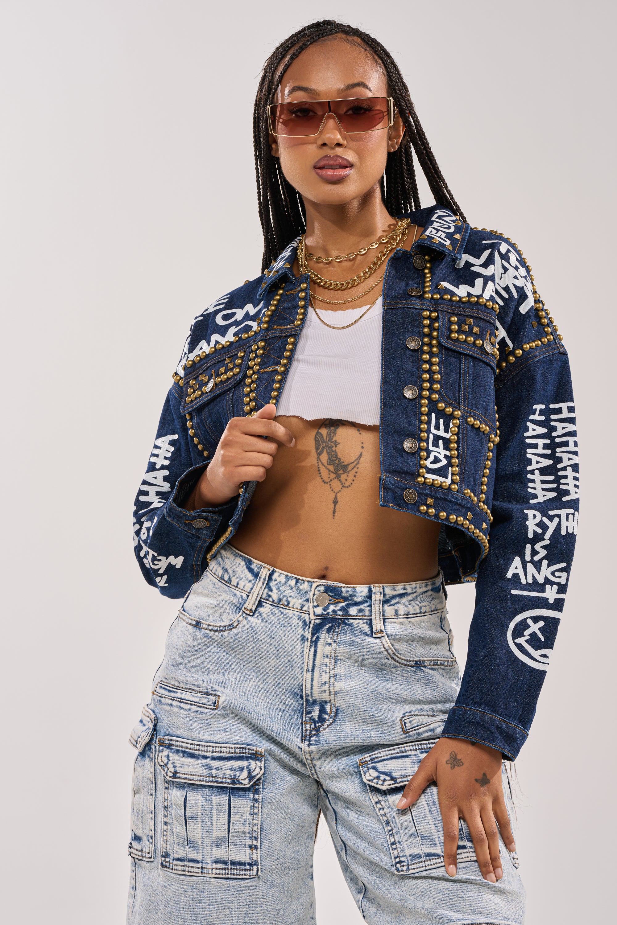 A stylish woman with braided hair poses confidently in the RHYTHM OF THE NIGHT DENIM JACKET IN DARK BLUE, featuring bold graffiti lettering, paired with a cropped white top, light blue cargo jeans, layered gold necklaces, and large sunglasses. 1
