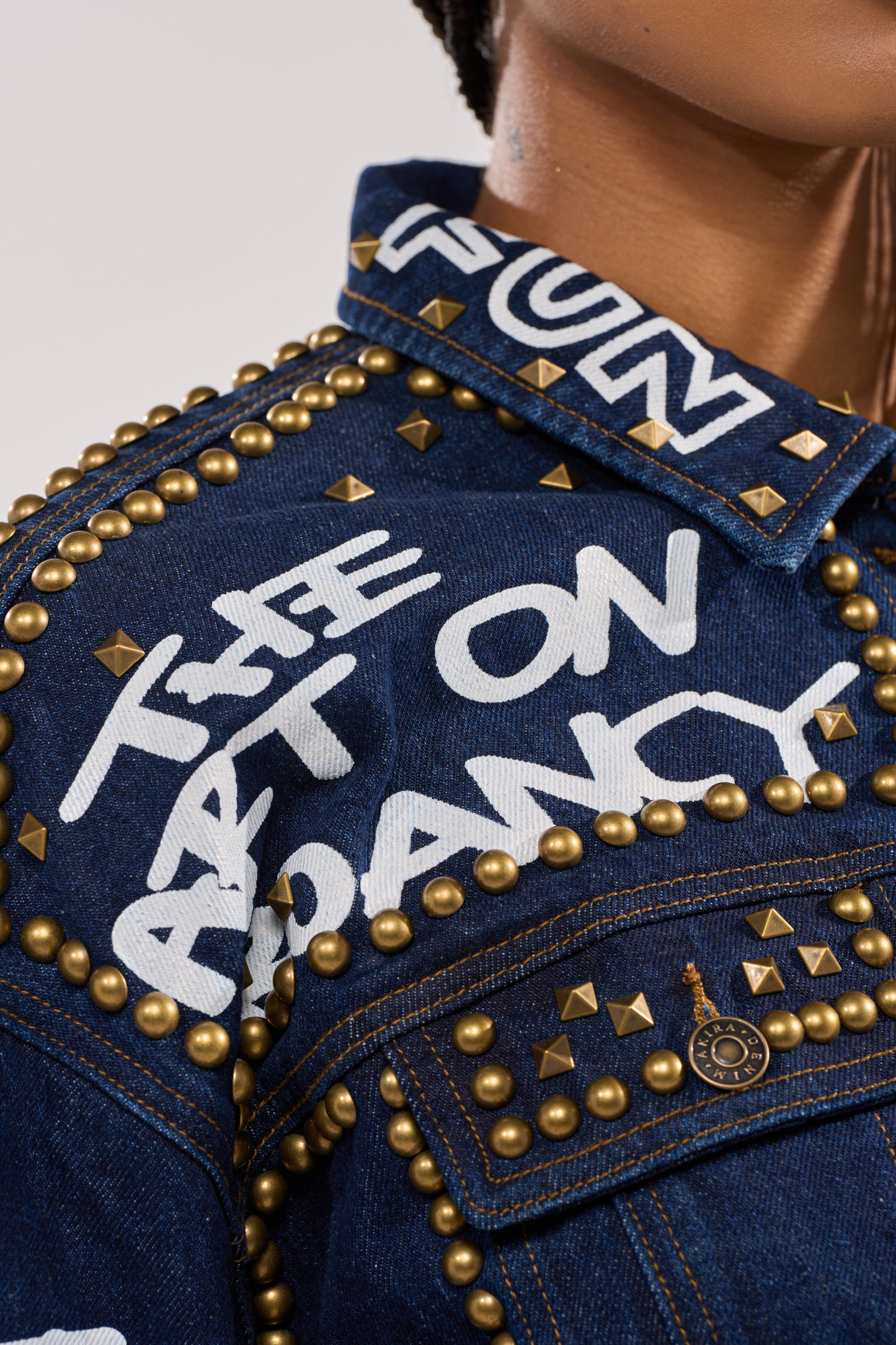 Close-up of the RHYTHM OF THE NIGHT DENIM JACKET IN DARK BLUE—a punk-inspired piece featuring gold studs, white graffiti lettering reading “USE ART ON FANCY,” decorative stitching, buttons, and part of a person's face and neck. 10