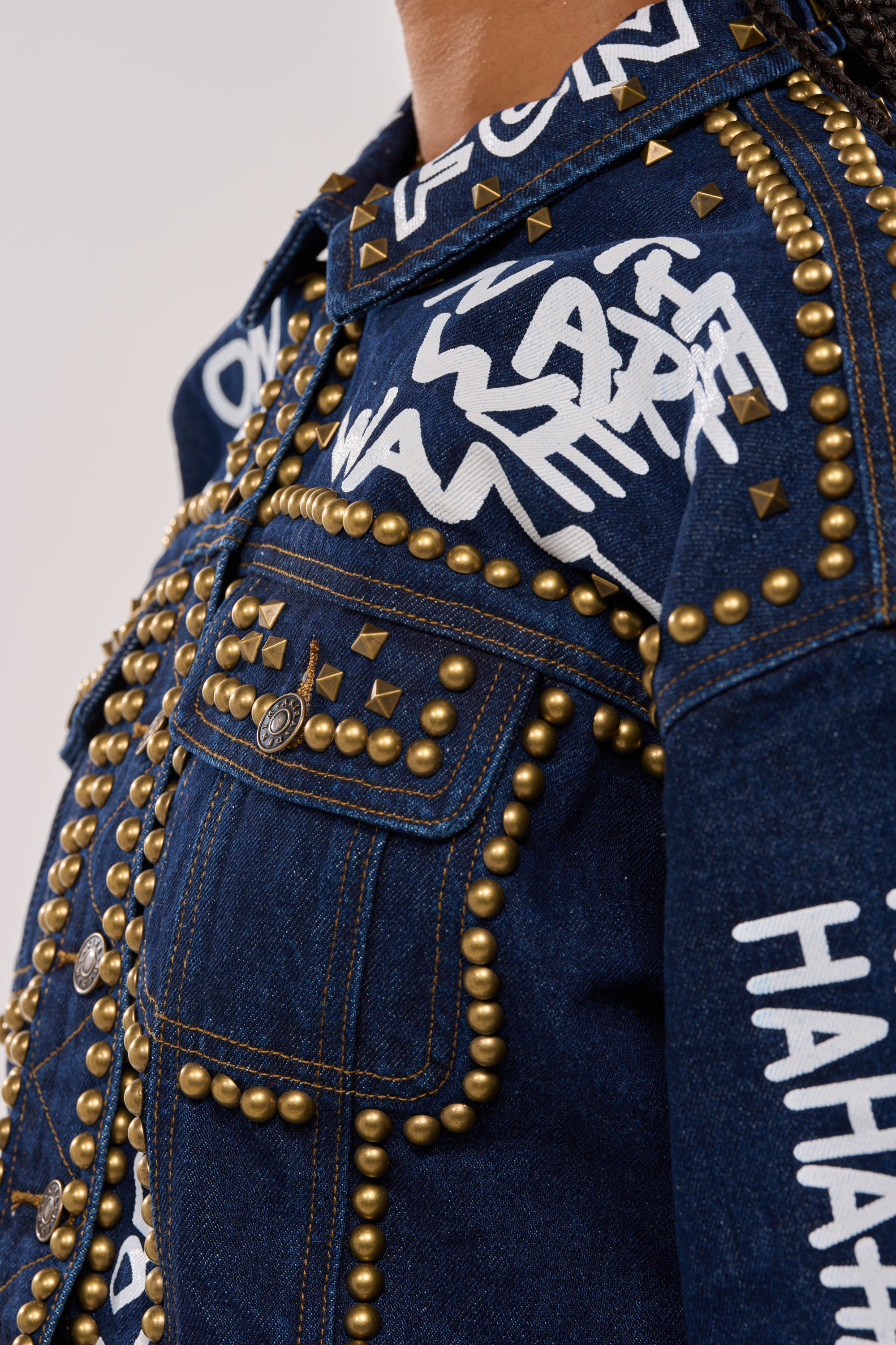 A close-up of the RHYTHM OF THE NIGHT DENIM JACKET IN DARK BLUE, featuring gold studs, white graffiti-style text like "HAHA," buttoned pockets, and abstract punk-inspired designs. 11