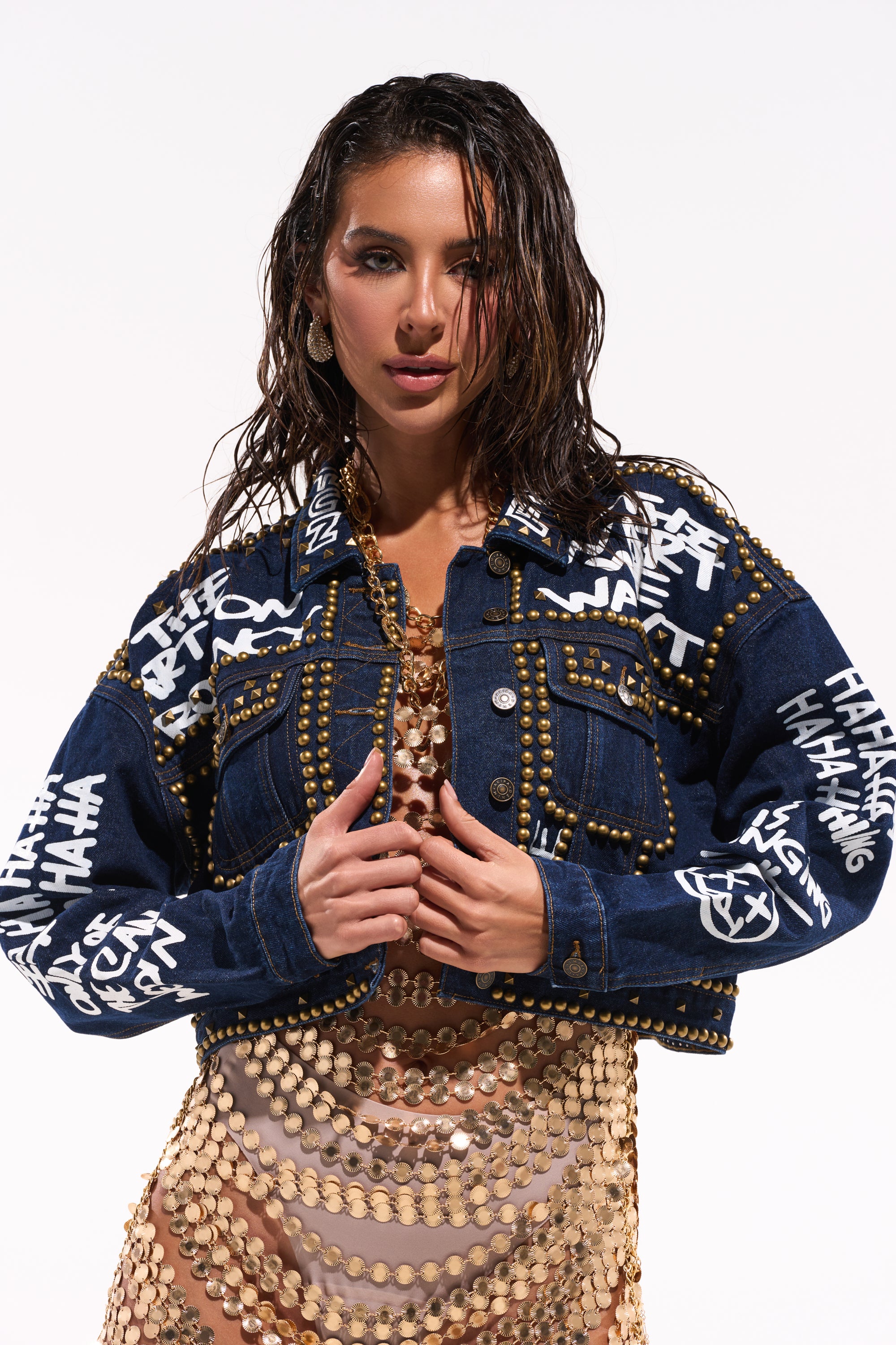 A woman with wet hair poses against a white background wearing the RHYTHM OF THE NIGHT DENIM JACKET IN DARK BLUE, a punk-inspired piece with white graffiti text and metallic studs, styled over a gold sequin mesh dress. 0