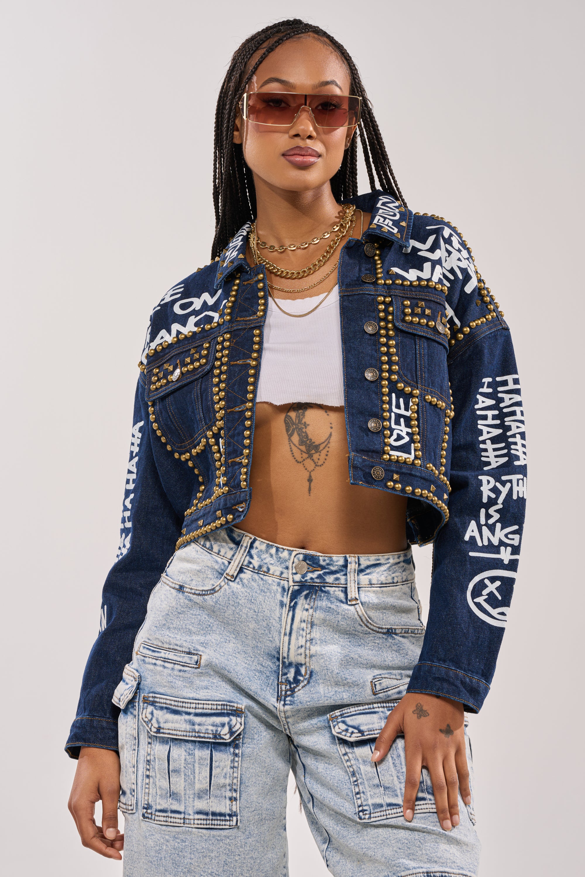 A stylish woman wears sunglasses, a white crop top, layered necklaces, high-waisted light wash cargo jeans with pockets, and the RHYTHM OF THE NIGHT DENIM JACKET IN DARK BLUE featuring punk-inspired graffiti lettering and studded details. 2