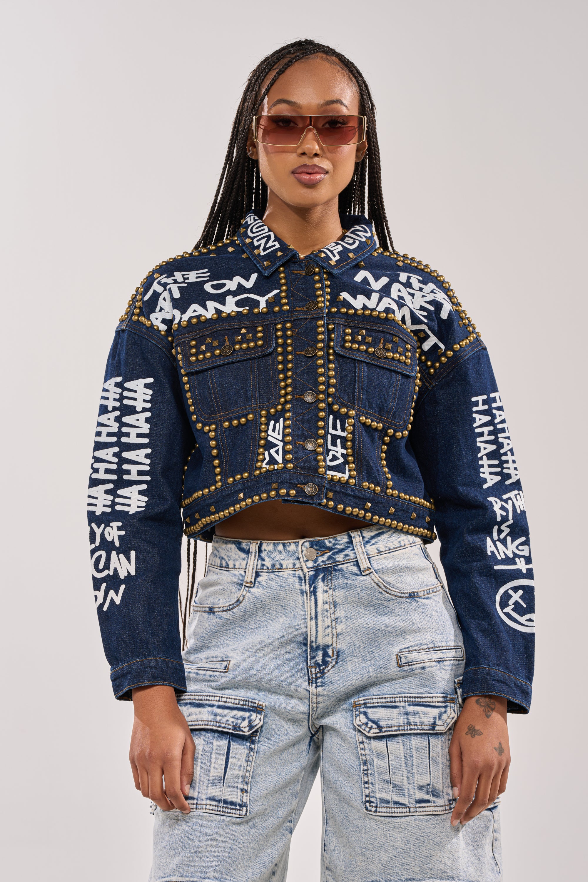 A woman with braided hair and sunglasses wears the RHYTHM OF THE NIGHT DENIM JACKET IN DARK BLUE with gold studs and white graffiti lettering, paired with light-wash jeans, standing confidently against a plain background. 3