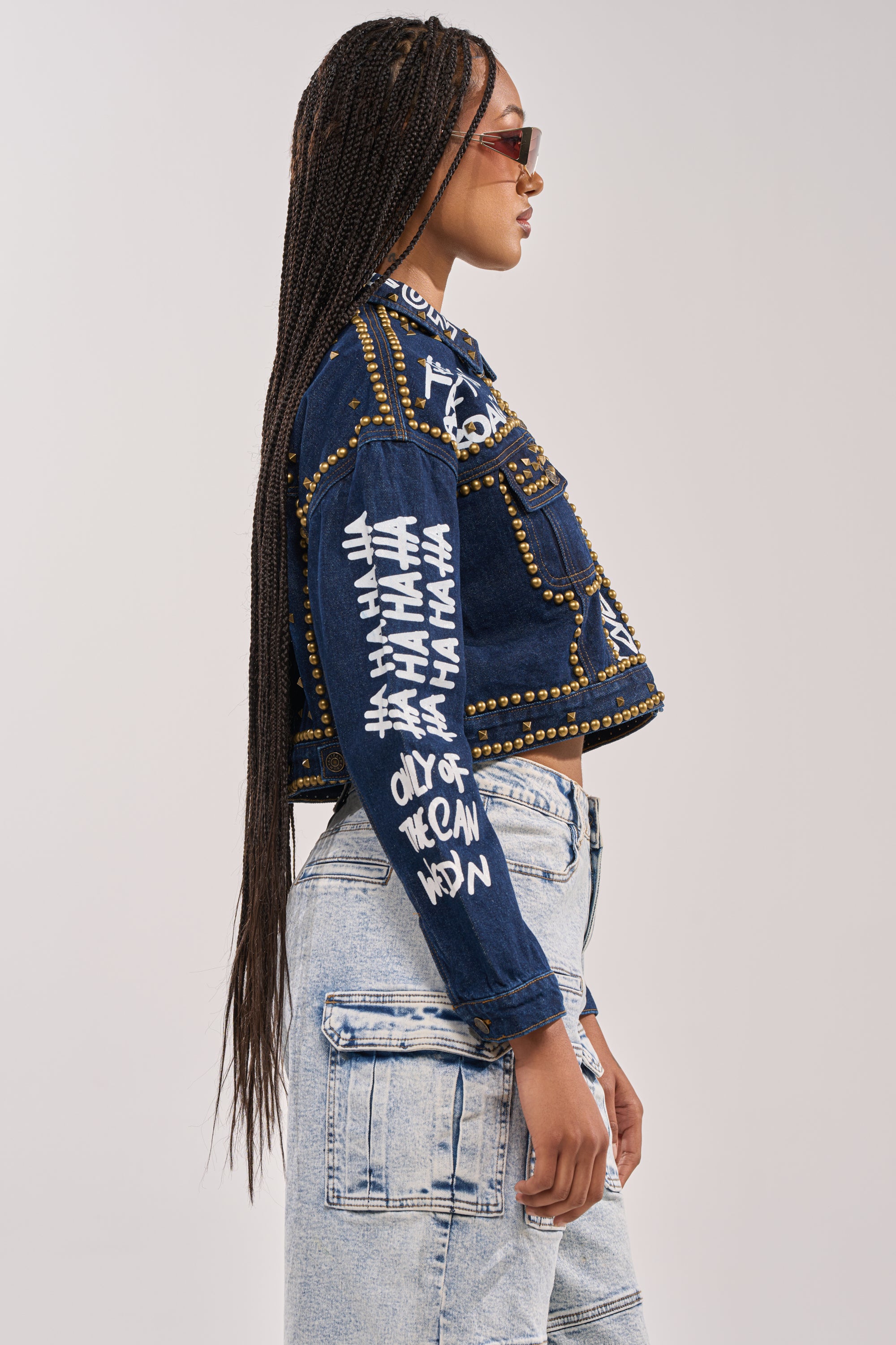 A woman with long braided hair and sunglasses poses in profile, wearing the RHYTHM OF THE NIGHT DENIM JACKET IN DARK BLUE with white graffiti-style text and light-wash cargo jeans against a plain background. 4