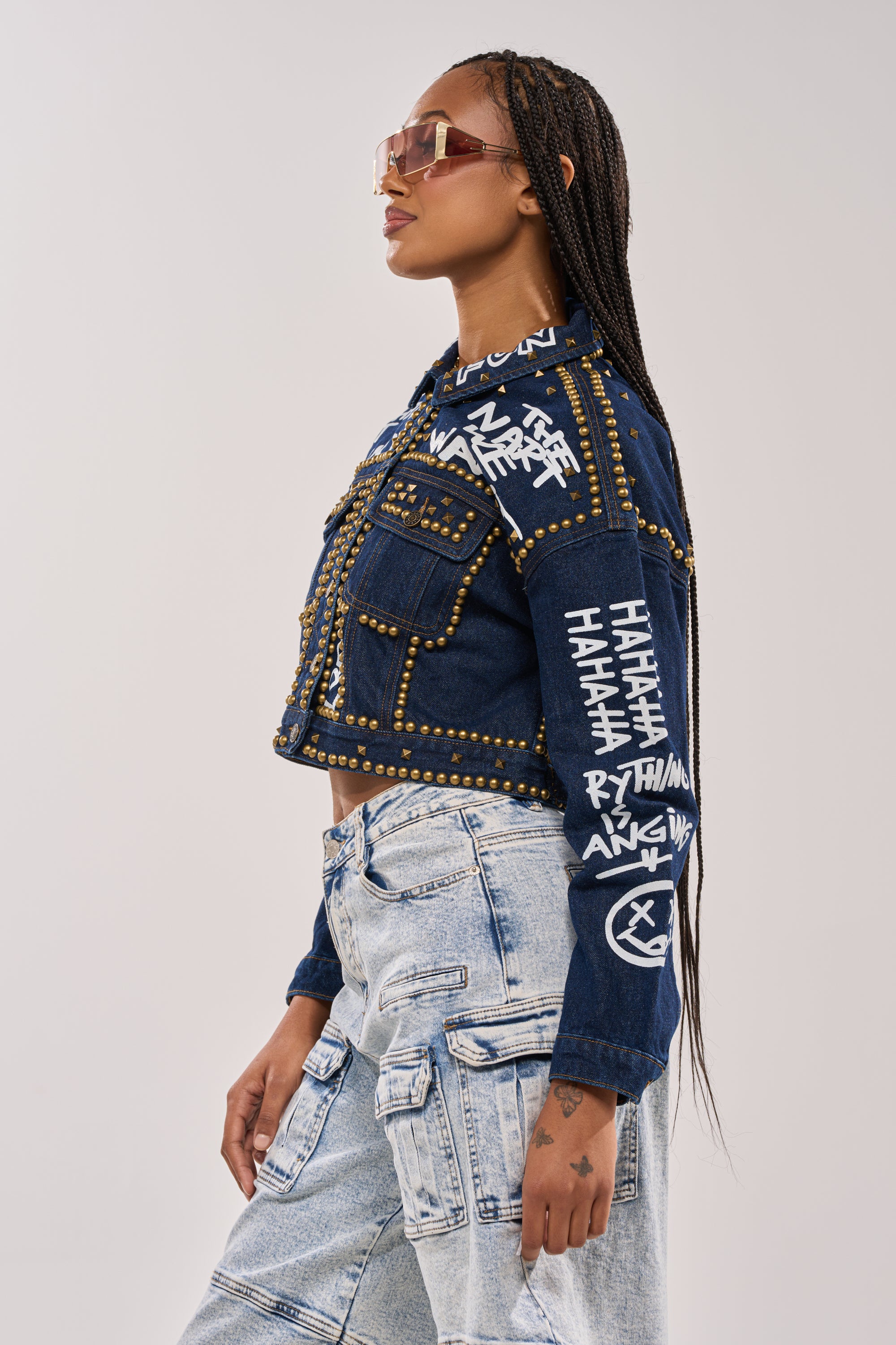 A woman with long braids and sunglasses poses in profile against a plain background, wearing the RHYTHM OF THE NIGHT DENIM JACKET IN DARK BLUE styled with light blue patchwork jeans. 5