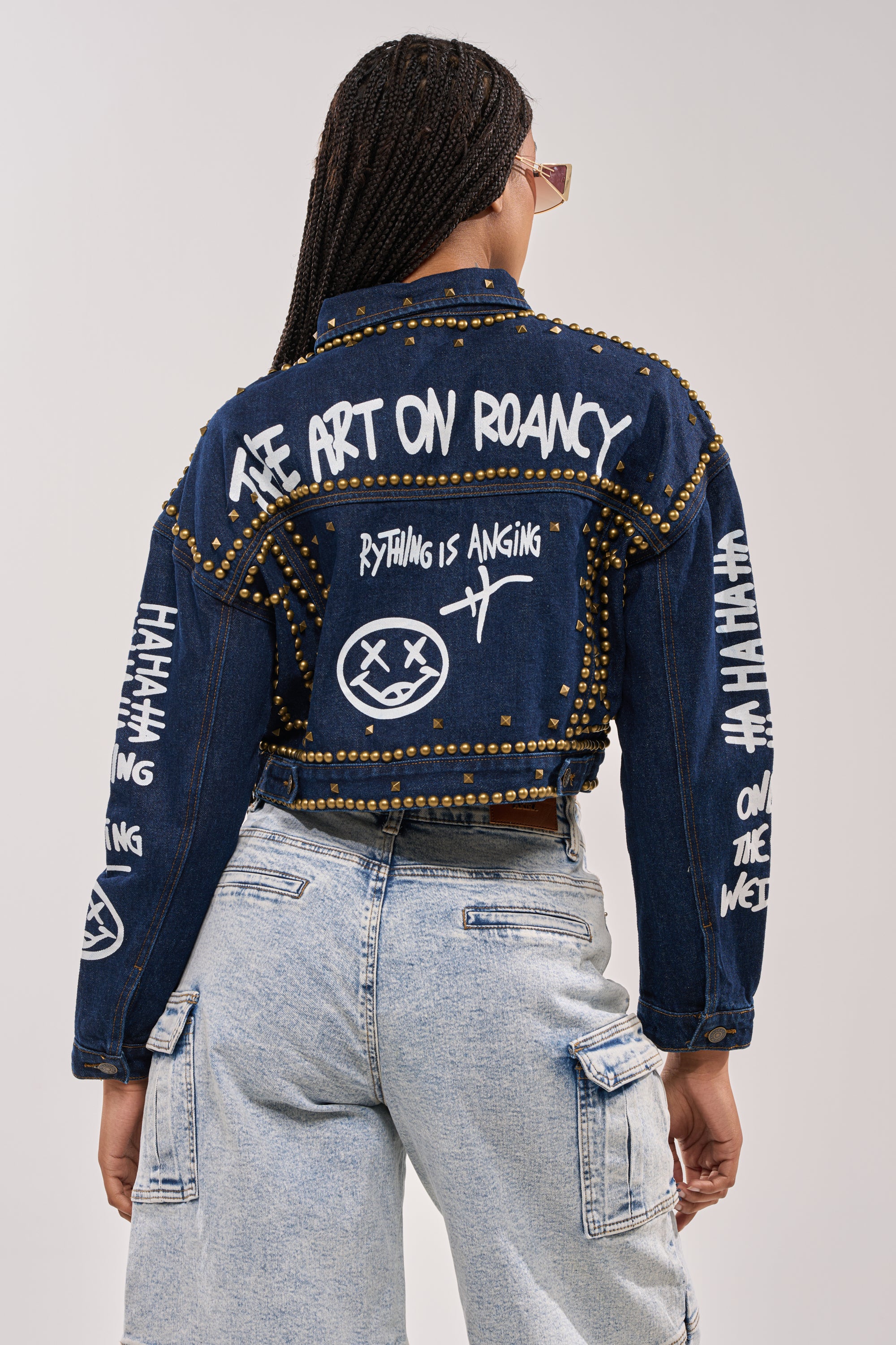 A person with braided hair faces away, wearing the RHYTHM OF THE NIGHT DENIM JACKET IN DARK BLUE with punk-style studs and graffiti lettering, paired with light blue cargo jeans. 6