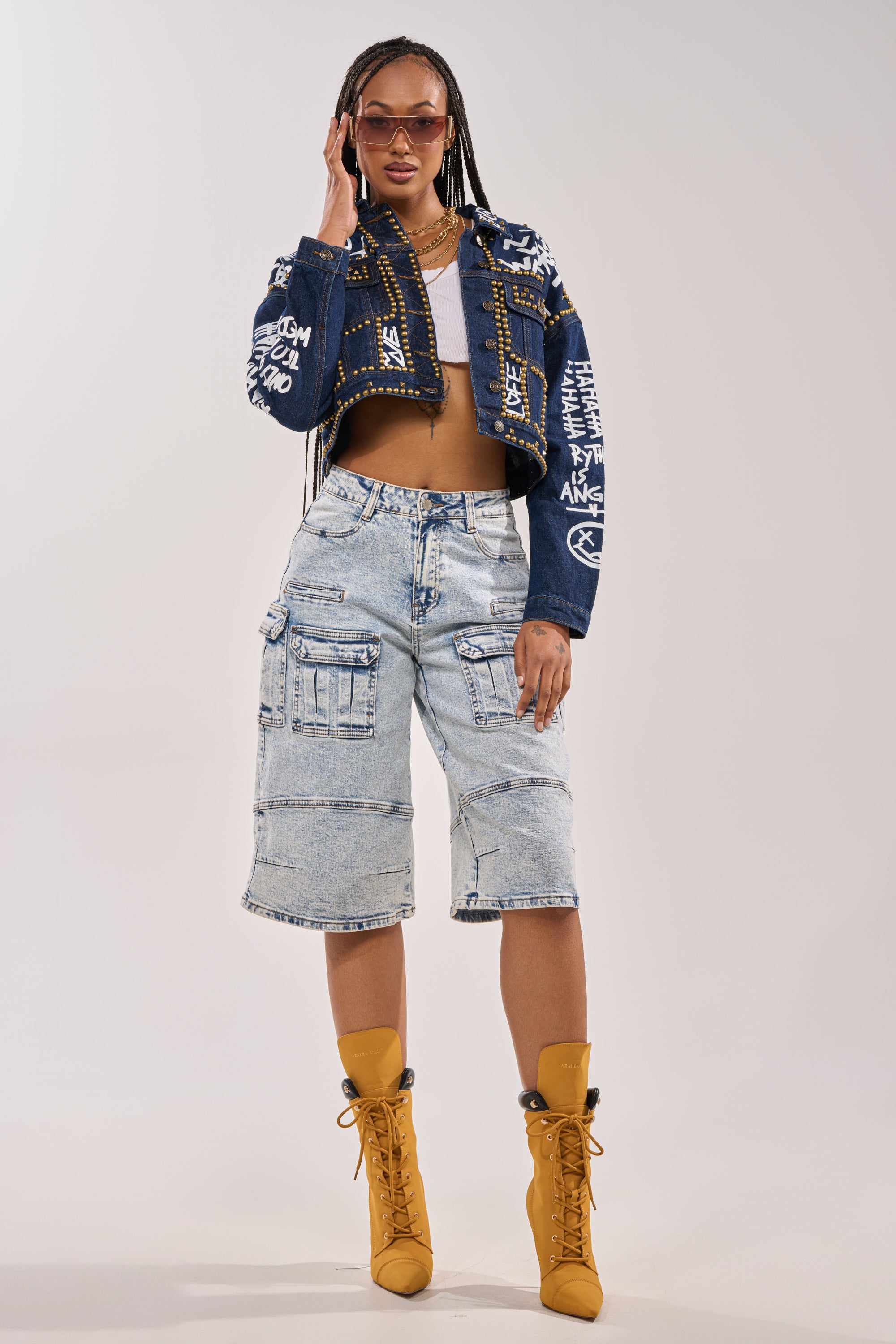 A woman poses confidently in the RHYTHM OF THE NIGHT DENIM JACKET IN DARK BLUE, graphic tee, light-wash denim cargo shorts, tan lace-up boots, sunglasses, and gold jewelry against a plain background. 7