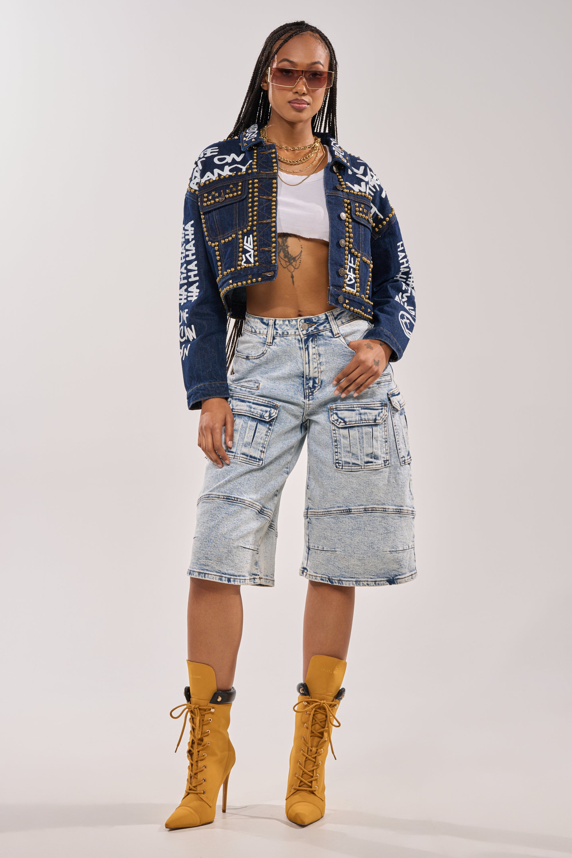 A woman with long braids wears sunglasses, layered necklaces, a white crop top, tan high-heeled boots, light-wash knee-length cargo shorts, and the RHYTHM OF THE NIGHT DENIM JACKET IN DARK BLUE, standing confidently against a plain background. 8
