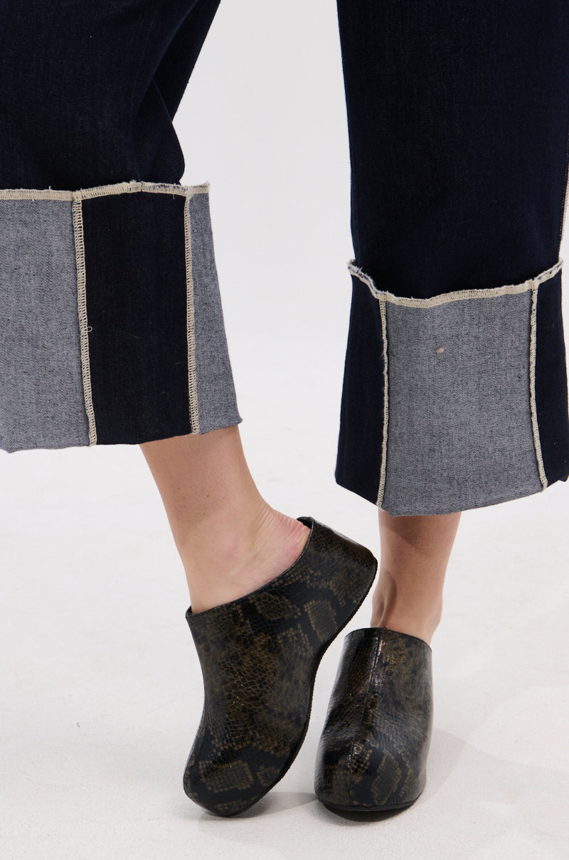 A person wears dark blue cuffed jeans with exposed seams and AZALEA WANG DARWIN CLOG IN BLACK SNAKE slip-ons, featuring Comfort Cushion insoles, standing on a white surface with one foot slightly raised and crossed in front.