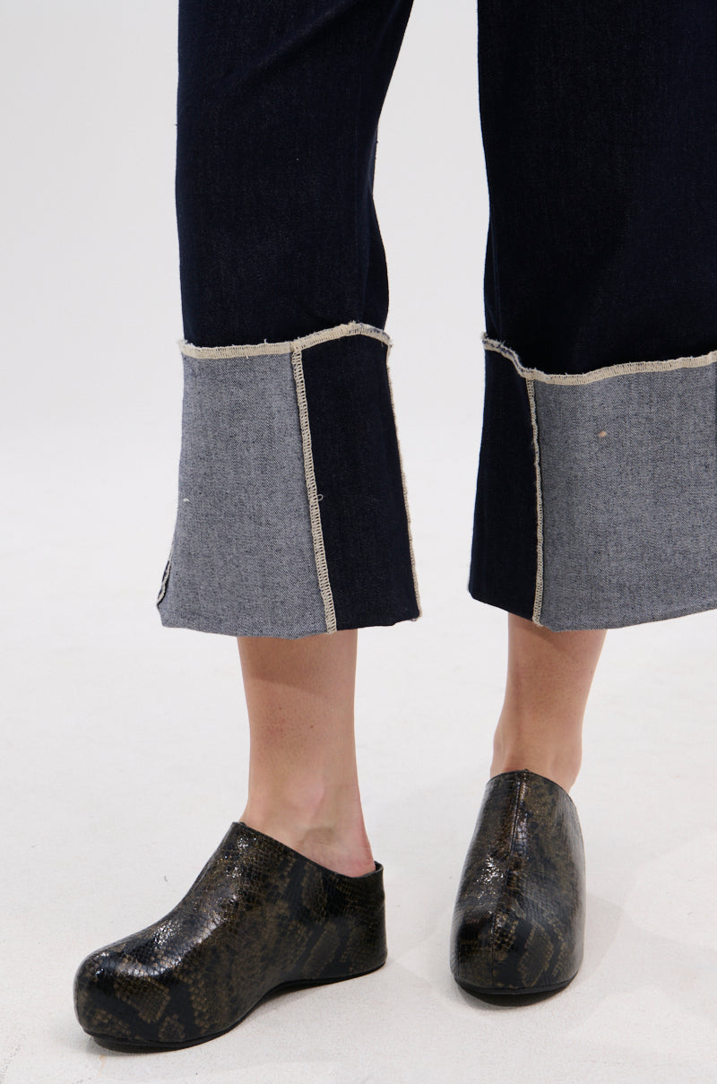 A person wearing dark denim pants with wide, light gray cuffs stands on a light floor, showing only their lower legs and feet in AZALEA WANG DARWIN CLOG IN BLACK SNAKE featuring a Comfort Cushion insole.