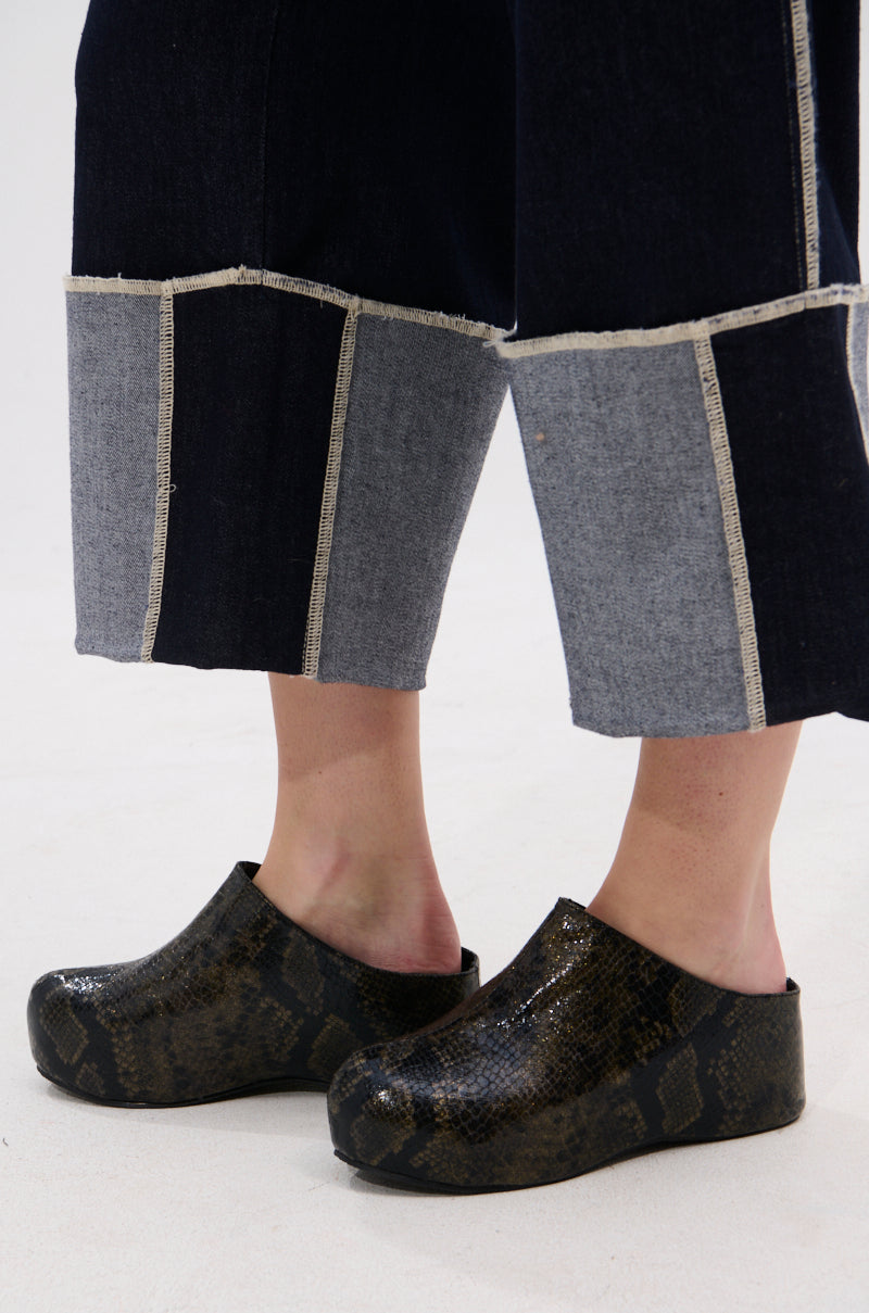 A person wears dark wide-leg pants with light gray patchwork hems and the AZALEA WANG DARWIN CLOG IN BLACK SNAKE, featuring a shiny snakeskin print and Comfort Cushion insole, while standing on a light surface.