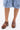 A person wearing knee-length light denim shorts with frayed hems and the AZALEA WANG DARWIN CLOG IN BROWN SNAKE faux leather clogs stands on a white background.