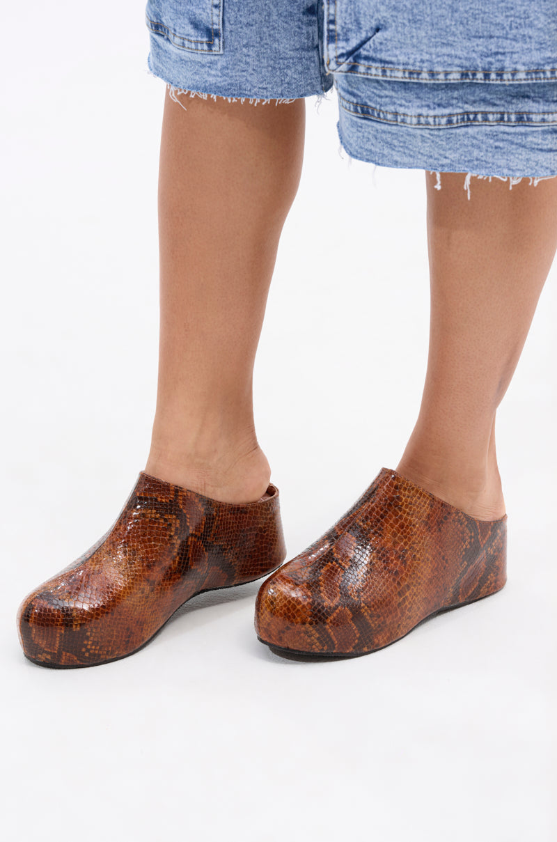 Person standing on a white background wearing blue denim shorts and AZALEA WANG DARWIN CLOG IN BROWN SNAKE, featuring a chunky heel.