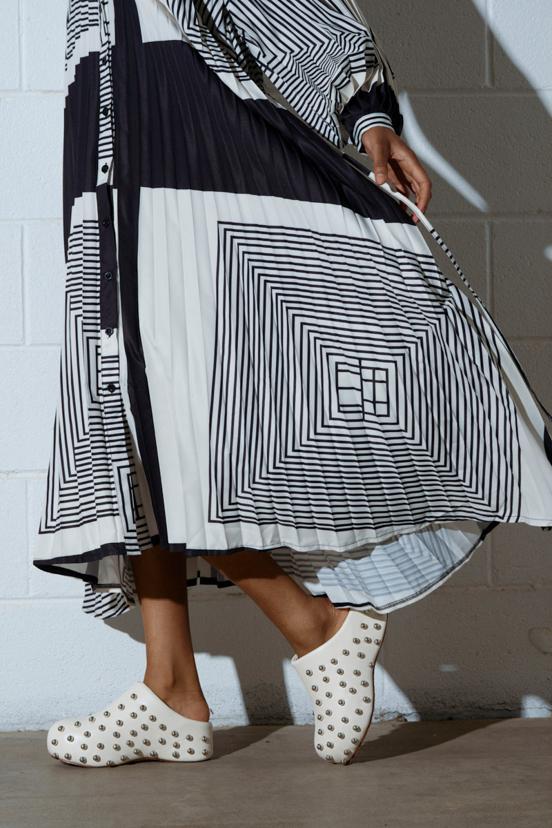 A person in a black and white geometric dress confidently walks past a white brick wall, wearing chic almond toe AZALEA WANG DARWIN STUD FLAT CLOGS IN BONE with metallic studs, adding flair to their look.