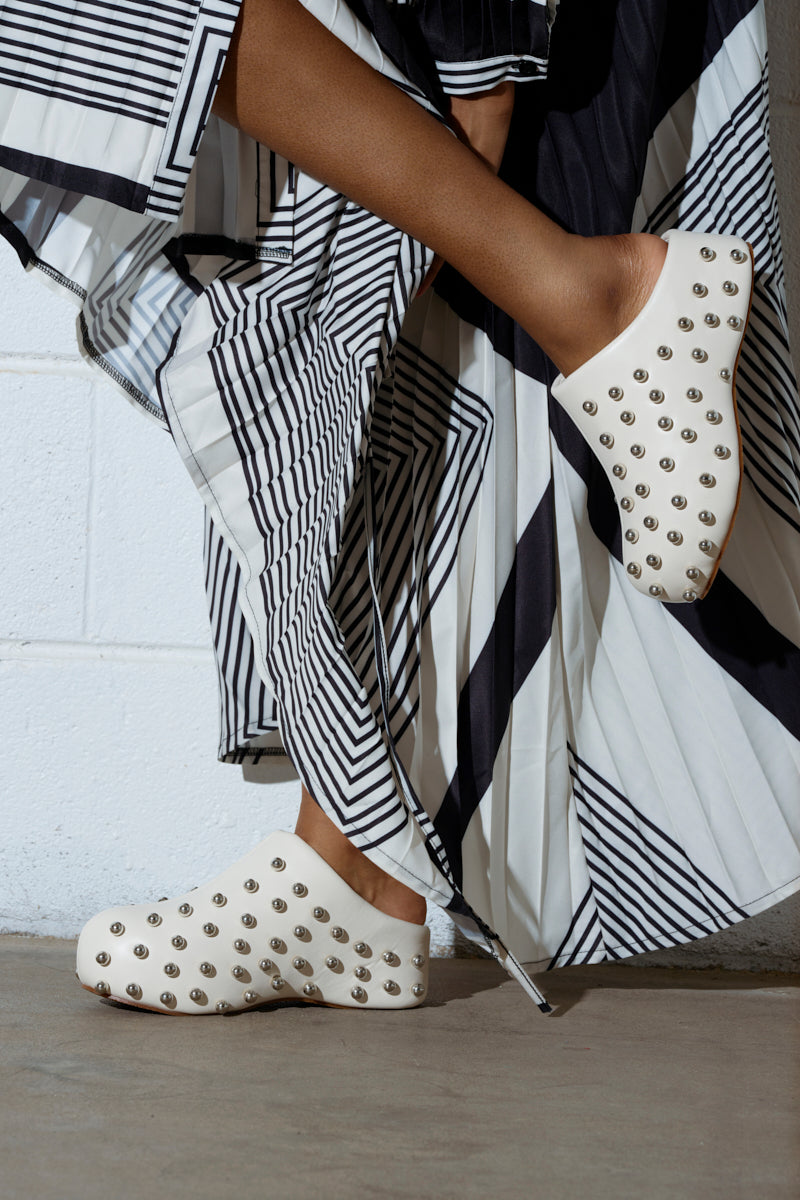 Wearing the AZALEA WANG DARWIN STUD FLAT CLOG IN BONE, a person stands against a white brick wall, showcasing the clogs' almond toe design and modern flatform sole beneath their black and white striped pleated skirt.