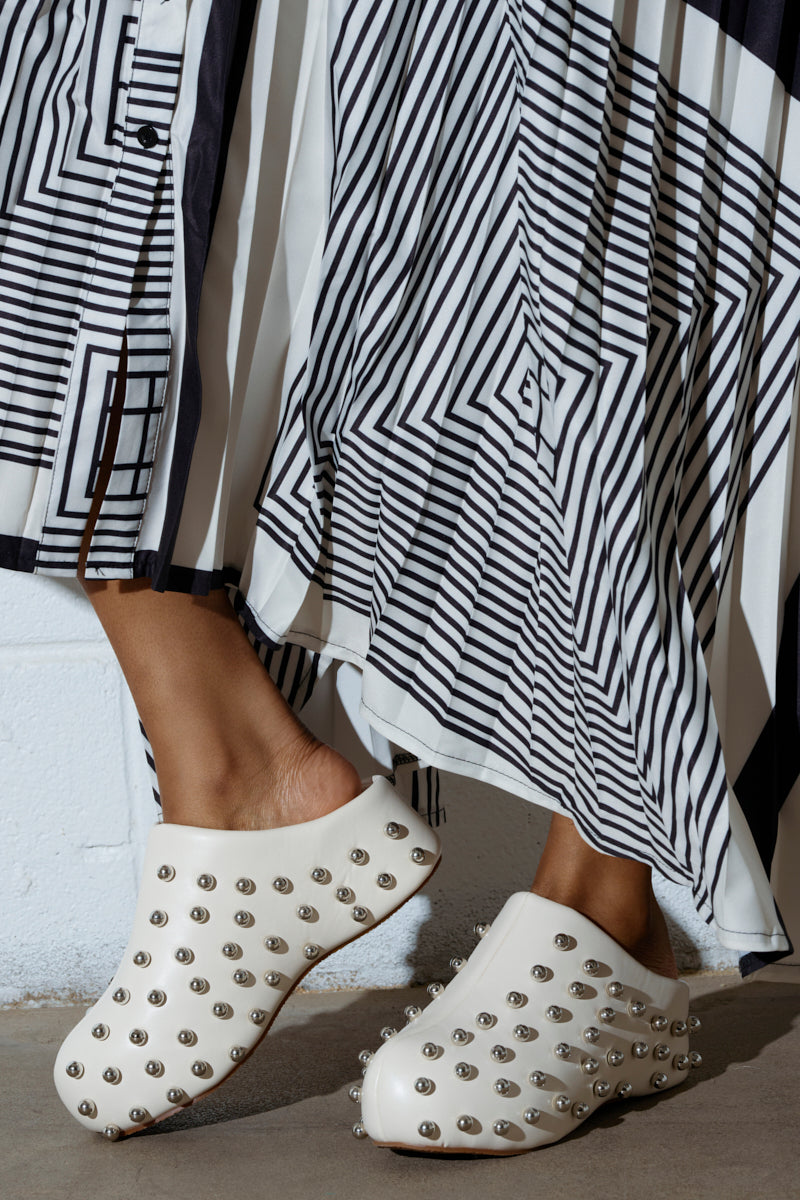 A person is seen wearing the AZALEA WANG DARWIN STUD FLAT CLOG IN BONE, crafted from faux leather with metal studs and an almond toe design. These white clogs feature a subtle flatform sole and are paired with a flowing black-and-white geometric skirt against a plain white wall backdrop.