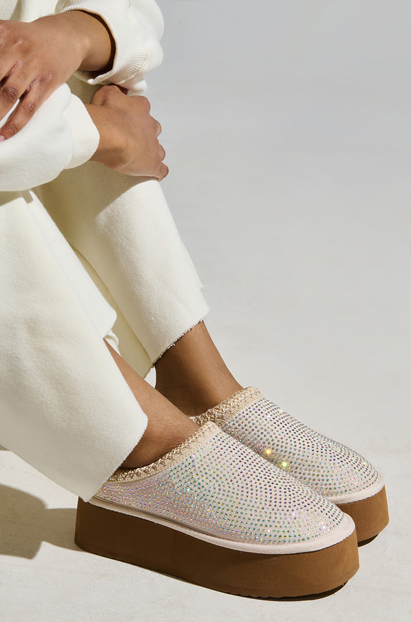 A person is seated, wearing white pants and AZALEA WANG DAVIAT WHITE EMBELLISHED SLIPPER FLATS with a brown sole. The rhinestone-embellished slippers reflect light beautifully against the simple, neutral gray background.