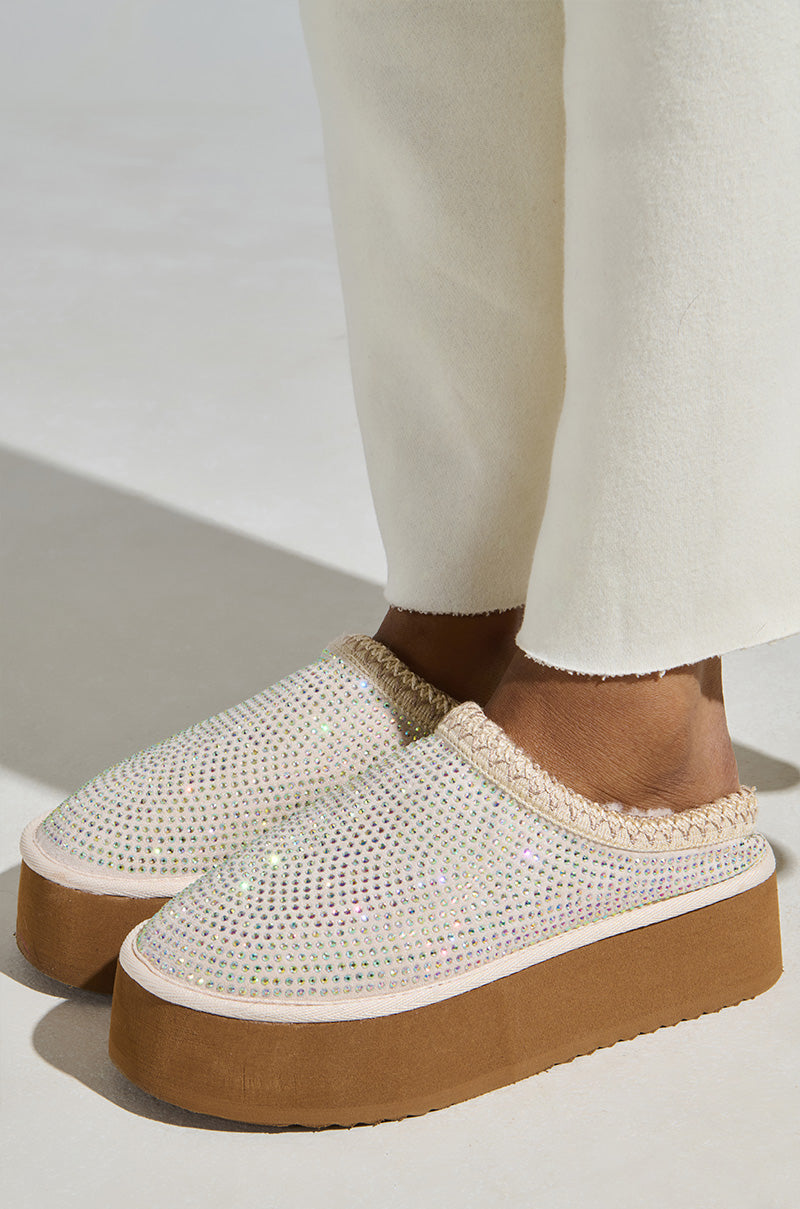 Standing on a white surface, a person wears cream-colored pants and shimmering AZALEA WANG DAVIAT WHITE EMBELLISHED SLIPPER FLATS with brown soles. The slippers' dazzling rhinestone designs add a touch of sparkle to the ensemble.