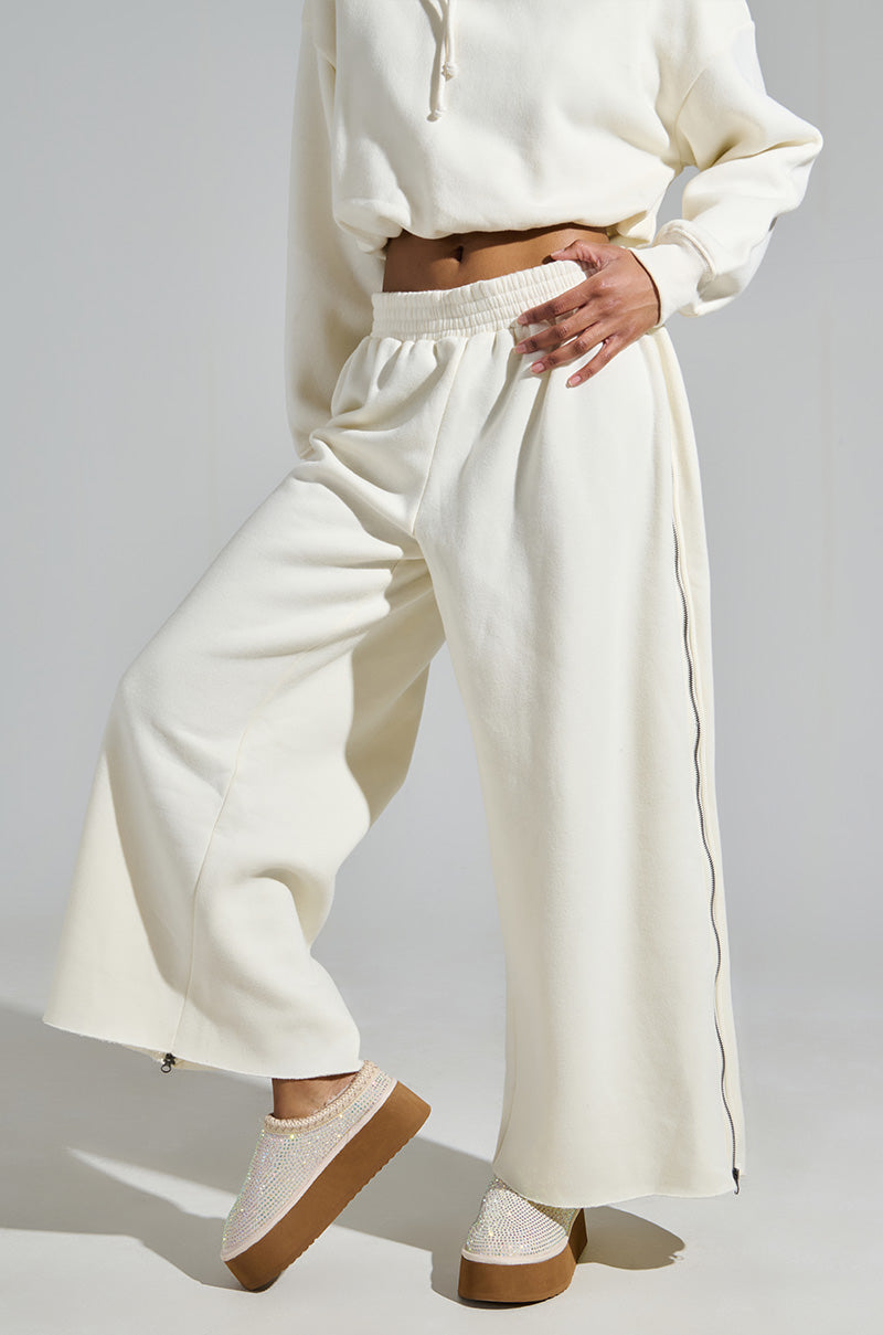 A person wearing a cream-colored hoodie and wide-leg pants stands against a neutral background, exuding confidence with their hand on their hip, and sporting AZALEA WANG DAVIAT WHITE EMBELLISHED SLIPPER FLATS adorned with rhinestones.