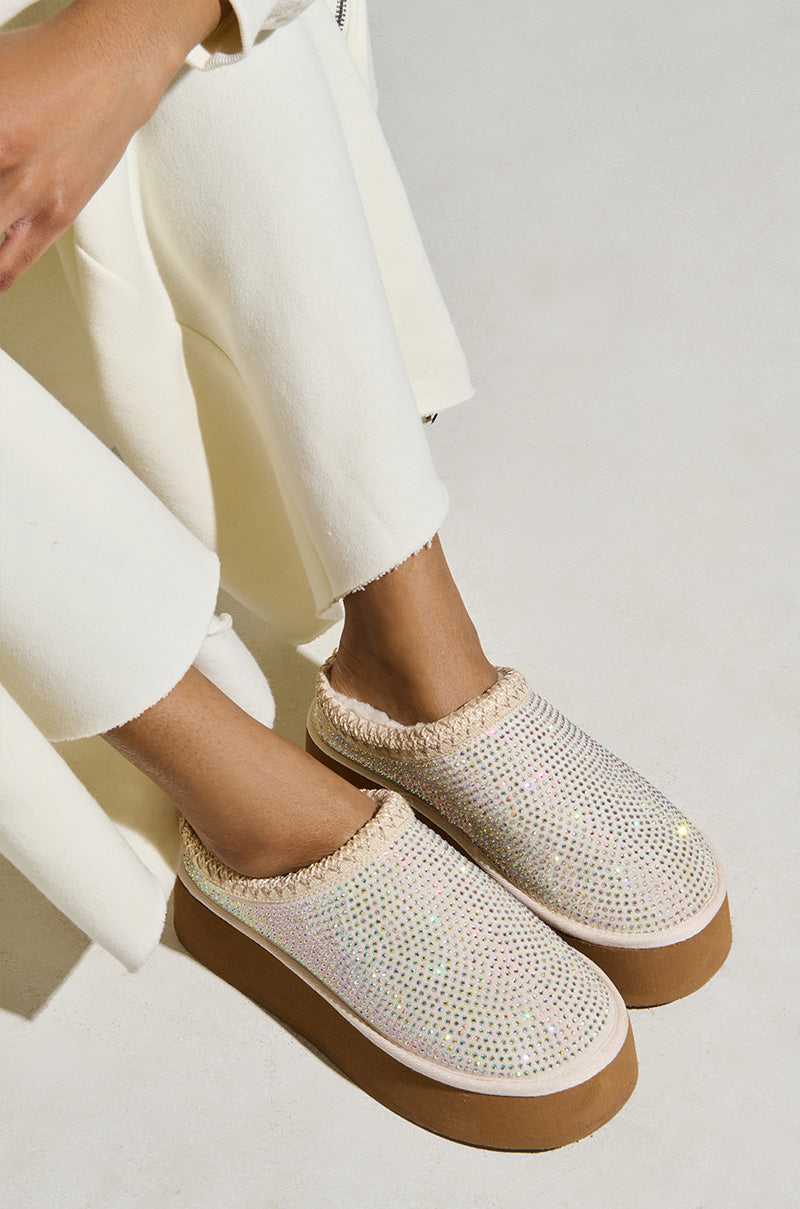 Someone is wearing cream-colored pants and the AZALEA WANG DAVIAT WHITE EMBELLISHED SLIPPER FLAT, which features glittery uppers and rhinestone-adorned thick brown soles. The eye-catching shimmer of these slippers stands out against the light, neutral backdrop as they sit with their feet crossed.