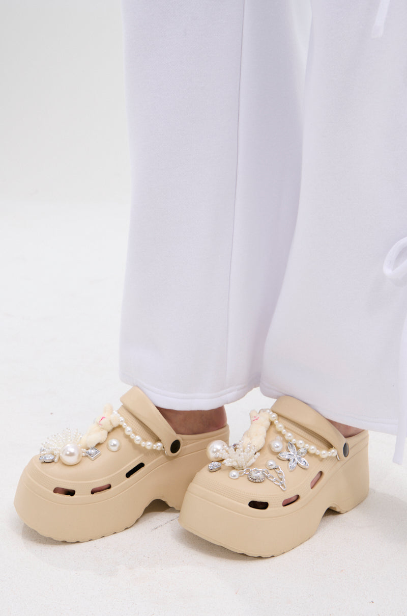 A person wearing loose white pants and AZALEA WANG DAVIES BEIGE EMBELLISHED FLATS, featuring pearls, rhinestones, and jewel-like accents, stands on a white surface.