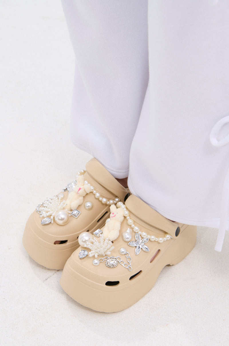 AZALEA WANG DAVIES BEIGE EMBELLISHED FLAT platform clogs, adorned with pearls, beads, jeweled charms, and a plush bunny charm, styled with loose white pants on a light-colored floor.