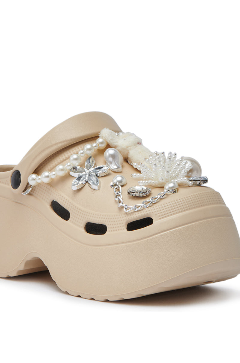 The AZALEA WANG DAVIES BEIGE EMBELLISHED FLAT is a beige platform clog featuring rhinestone accents, faux pearls, a silver flower, metallic chain, and a white fuzzy accessory, shown against a white background.