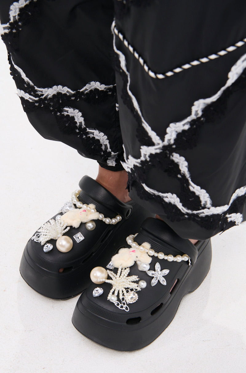 Someone wearing AZALEA WANG DAVIES BLACK PEARL RHINESTONE FLAT clogs with black pants featuring white embroidered patterns.