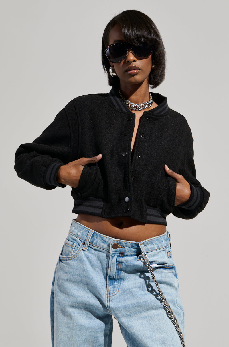 A person wearing the DAVIS EVERYDAY CROPPED VARSITY BOMBER IN BLACK, light blue jeans, and sunglasses, styled with a chain necklace. They are posing confidently against a plain background.