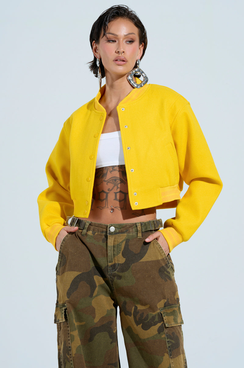 A person donning the DAVIS EVERYDAY VARSITY BOMBER IN YELLOW with a cropped silhouette over a white top, showcasing their tattooed midriff. They accessorize with large circular earrings and camouflage cargo pants, standing against a light background. 0