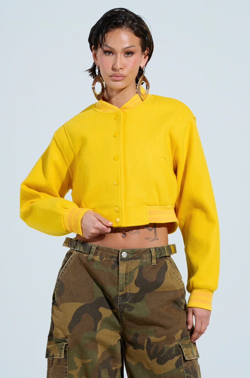 A person stands confidently in the DAVIS Everyday Varsity Bomber in yellow with button details and camo cargo pants. The jacket's ribbed knit collar adds a touch of flair. They have short dark hair and are wearing large, dangling earrings, set against a simple, light blue background. 1