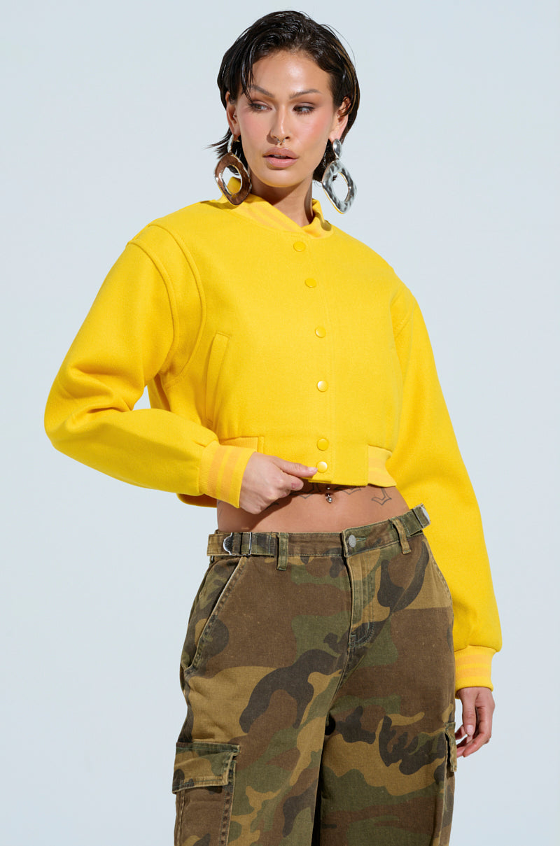 A person wearing the Davis Everyday Varsity Bomber in yellow, featuring a ribbed knit collar, confidently poses with one hand on their hip and is styled in camouflage pants. Their wet-look hair and statement earrings stand out against the plain background, enhancing the flair of their cropped ensemble. 2