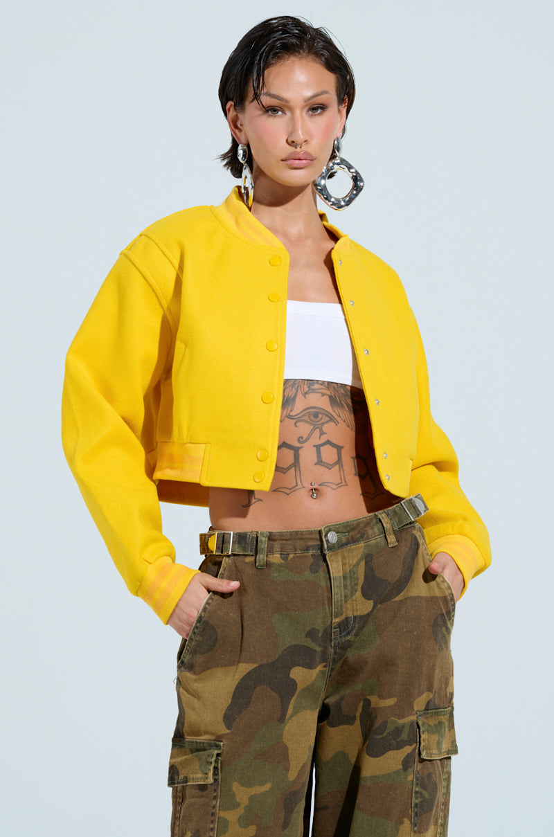 A person is dressed in the DAVIS EVERYDAY VARSITY BOMBER IN YELLOW, paired with a white top and adorned with large hoop earrings. They are also wearing camo-style cargo pants, revealing tattoos on their torso while their hands rest in their pockets. The background is light blue. 3