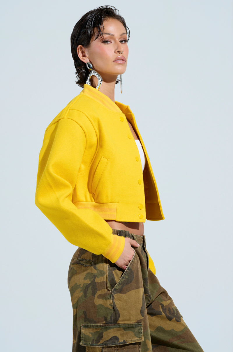 A person wearing the eye-catching Davis Everyday Varsity Bomber in Yellow, complete with a ribbed knit collar, along with camouflage cargo pants, strikes a confident pose against a light blue background. With short dark hair and large hoop earrings, they create a bold and stylish silhouette. 4