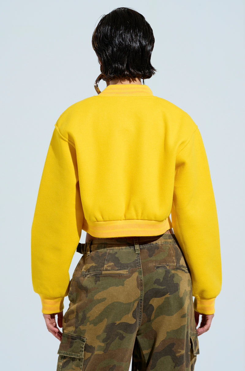 A person with short dark hair is seen from behind, wearing the vibrant yellow Davis Everyday Varsity Bomber, paired with camouflage cargo pants. The background is a plain light blue. 5