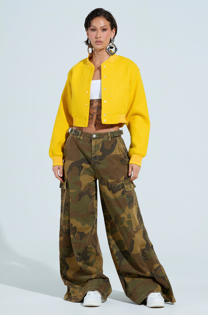A person with short hair is confidently standing against a light background, wearing the DAVIS Everyday Varsity Bomber in yellow, which features a ribbed knit collar. They have paired it with a white top, camouflage cargo pants, and white sneakers. Large earrings serve as bold accessories to complete the look. 6