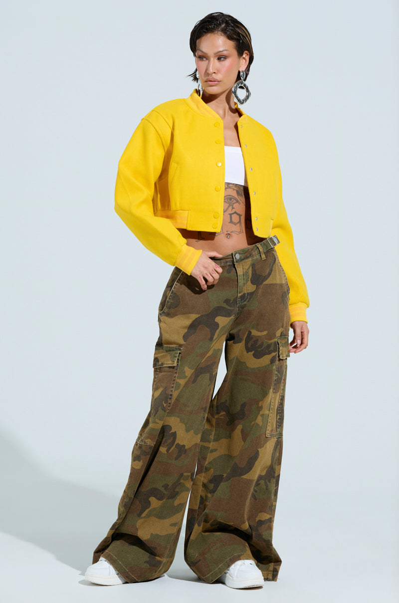 A person exudes confidence in the DAVIS EVERYDAY VARSITY BOMBER IN YELLOW, featuring a ribbed knit collar over a white top. They pair it with camo cargo pants and white sneakers, sporting short dark hair and accessorizing with large earrings, all set against a simple, light-colored background. 7