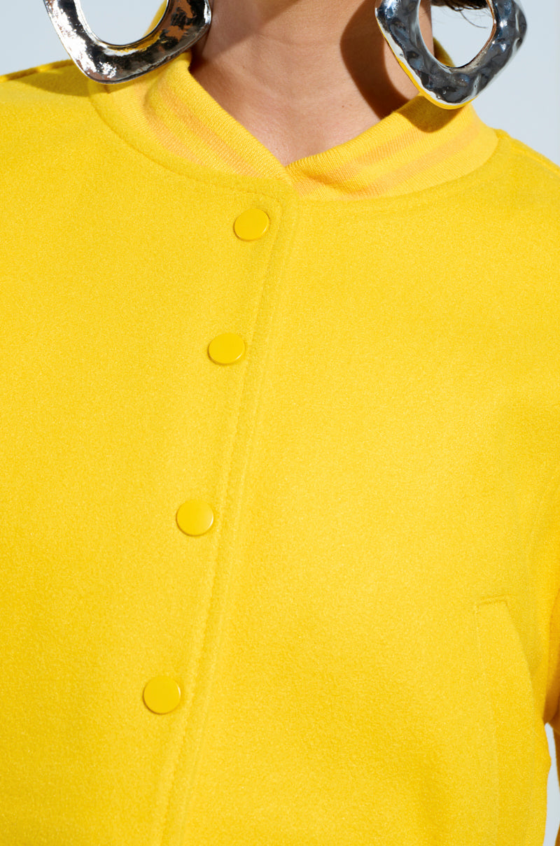 Close-up of a person wearing the DAVIS Everyday Varsity Bomber in Yellow, featuring large button details and a ribbed knit collar. They complement their look with shiny silver hoop earrings. The light background enhances the vivid color of this stylish ensemble. 8