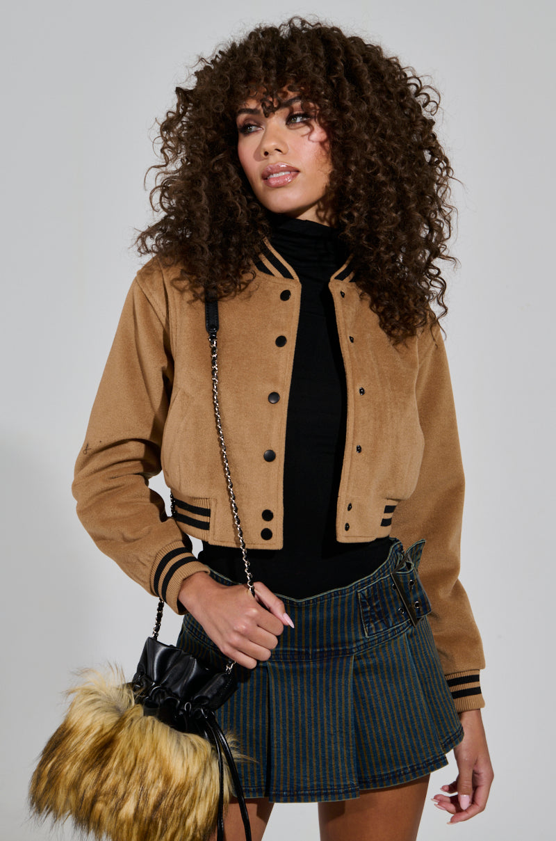 Wearing the DAVIE EVERYDAY BOMBER over a black turtleneck, paired with a striped skirt, and holding a handbag with a fur detail, a person with curly hair poses against a plain background.
