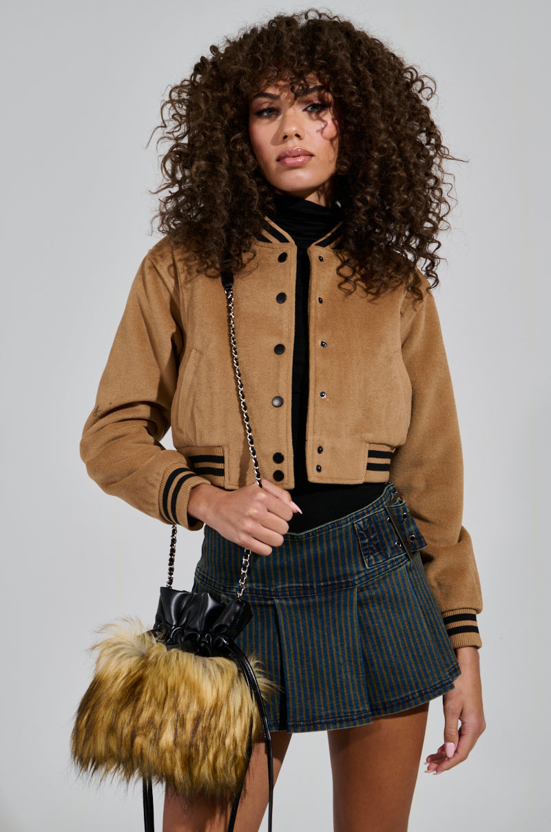 A person with curly hair wearing the DAVIE EVERYDAY BOMBER and a striped skirt poses confidently. They are holding a fuzzy teddy knit bag with a chain strap against a plain, light-colored background.