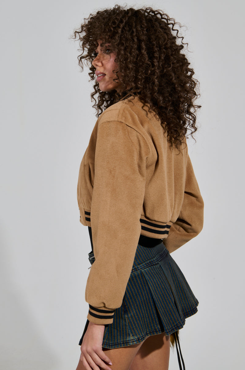 A person with curly hair is wearing the DAVIE EVERYDAY BOMBER in light brown, paired with a short, pleated skirt. The cropped silhouette of the jacket adds a modern touch as they stand sideways with a neutral expression against a plain background.