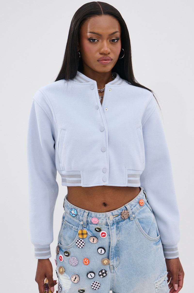 A woman wears the DAVIS EVERYDAY VARSITY BOMBER IN LIGHT BLUE, styled with high-waisted denim jeans decorated with colorful pins and patches, standing against a plain white background. 3