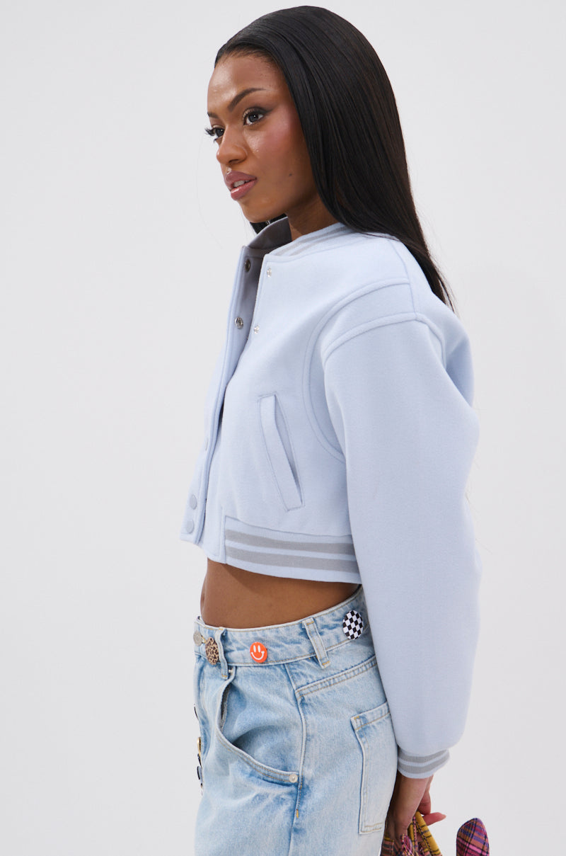 A woman with long straight hair stands in profile against a white background, wearing the DAVIS EVERYDAY VARSITY BOMBER IN LIGHT BLUE with a cropped fit and light-wash jeans, holding a small item in her right hand. 4