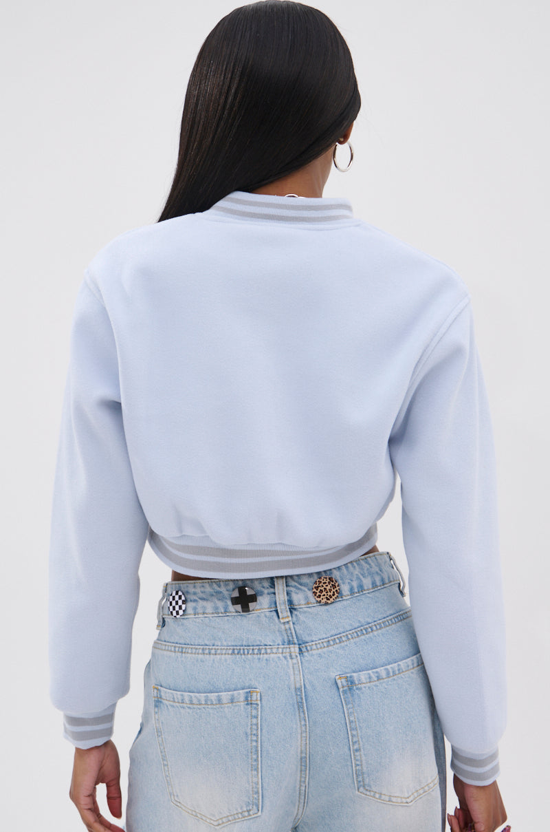 A person with long dark hair is shown from the back, wearing the DAVIS EVERYDAY VARSITY BOMBER IN LIGHT BLUE in a cropped style paired with high-waisted light wash jeans featuring decorative buttons on the waistband. 5