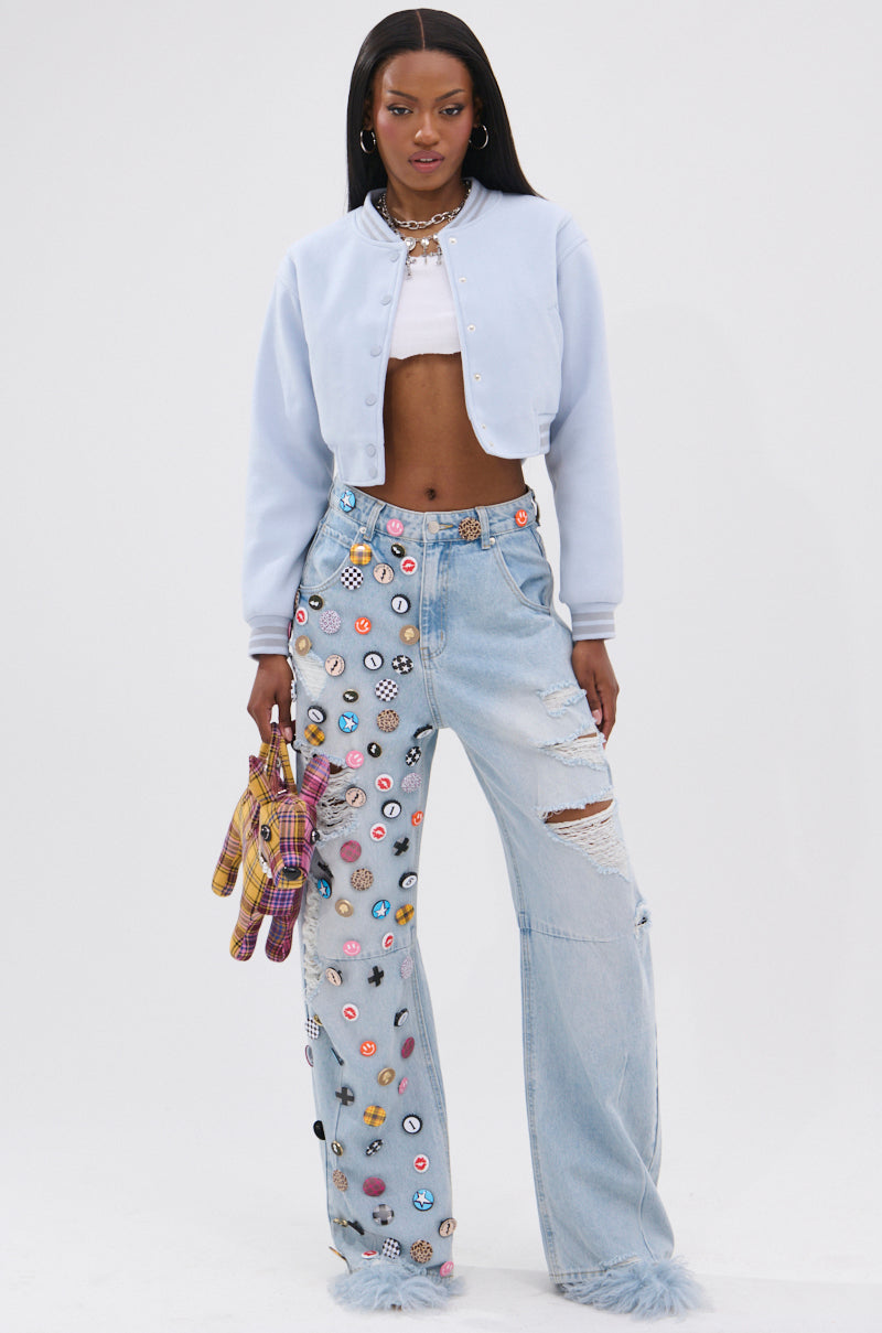 A woman stands confidently in the DAVIS EVERYDAY VARSITY BOMBER IN LIGHT BLUE, styled over a white crop top and light blue ripped jeans with colorful patches, fluffy ankle cuffs, and holding a plaid plush toy. 6