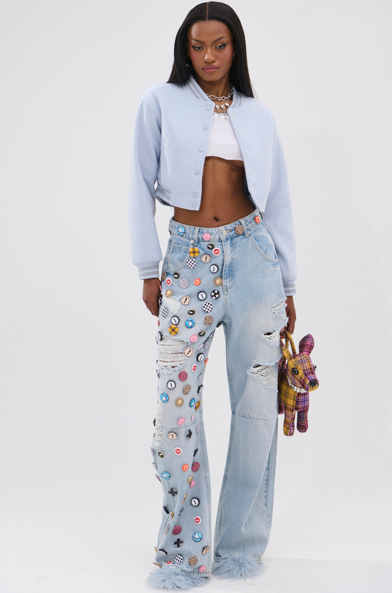 A woman with long straight hair and a confident look wears the DAVIS EVERYDAY VARSITY BOMBER IN LIGHT BLUE, a white crop top, patchwork distressed light blue jeans, and holds a small plaid stuffed dog. 7