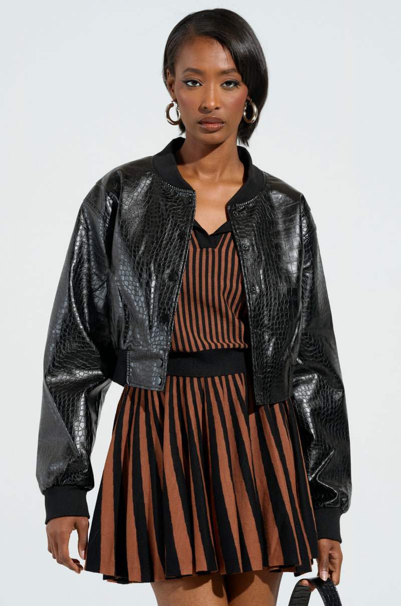 A person wearing the "WHAT I WANT FAUX LEATHER BOMBER" jacket over a brown and black striped dress stands against a light gray background. They have long hair, accessorized with hoop earrings, and are holding an embossed snakeskin pattern handbag.