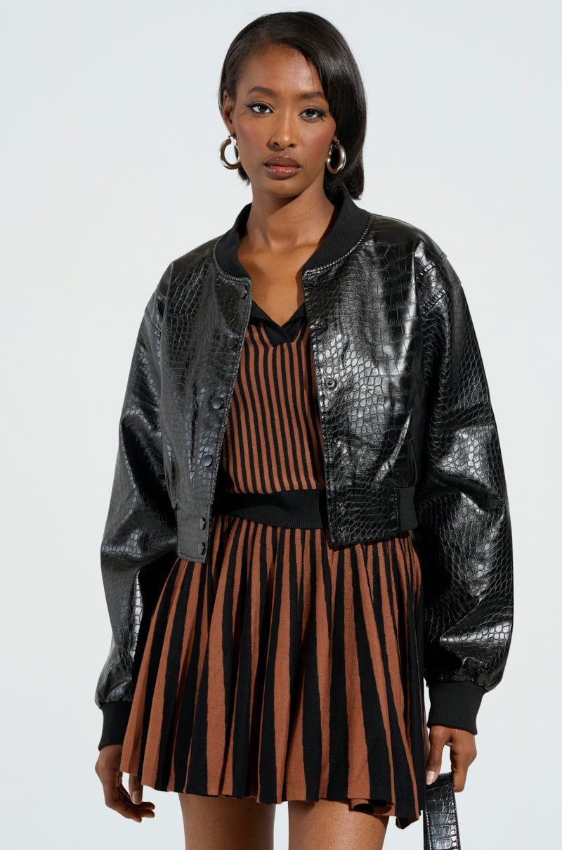 A person wearing the WHAT I WANT FAUX LEATHER BOMBER, which features a black faux leather material with a cropped silhouette, over a brown and black striped dress. They have long hair and are accessorized with hoop earrings, standing against a light background.